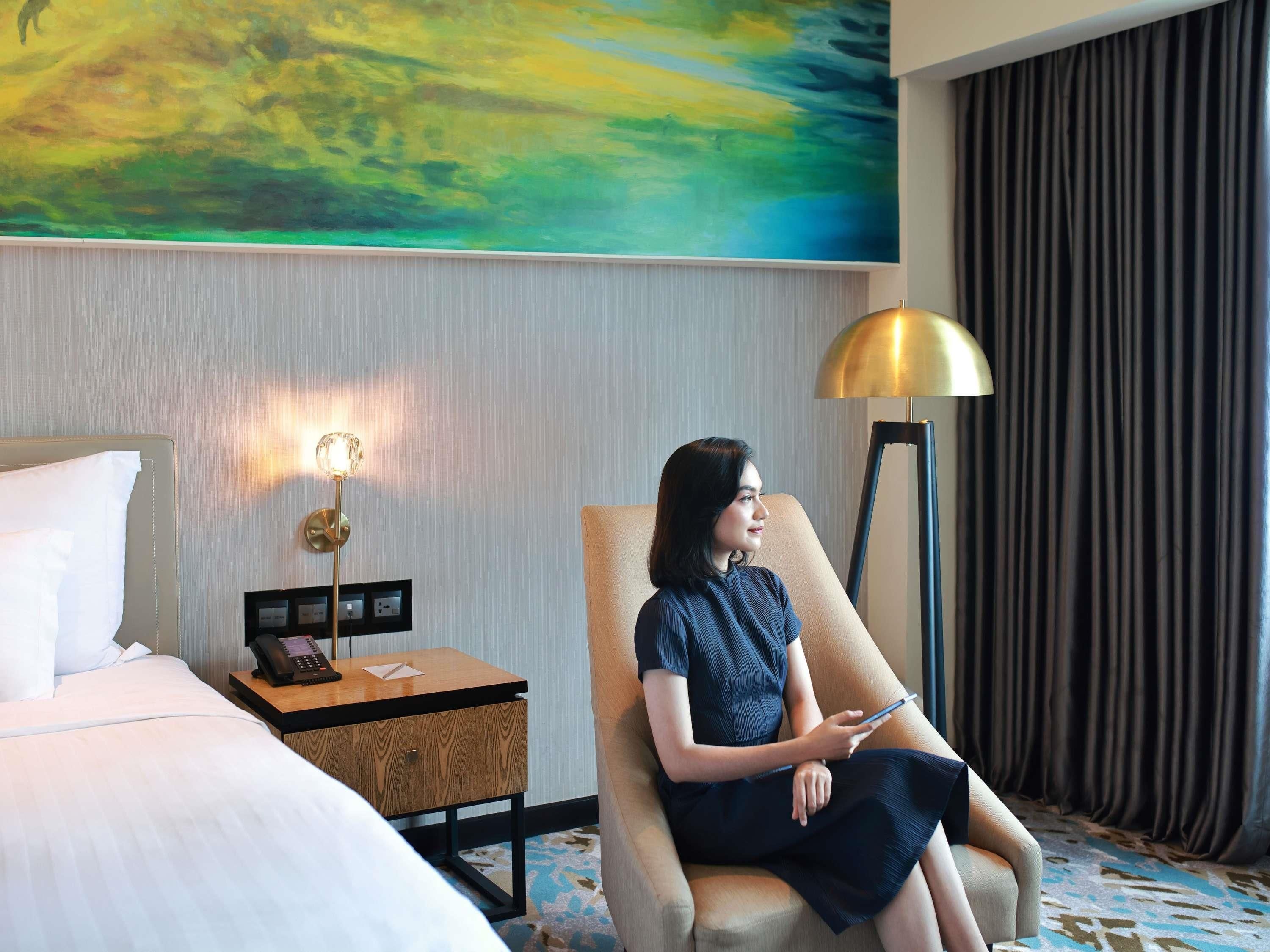 A hotel room with a bed, a desk with a lamp, a chair, and a painting on the wall.