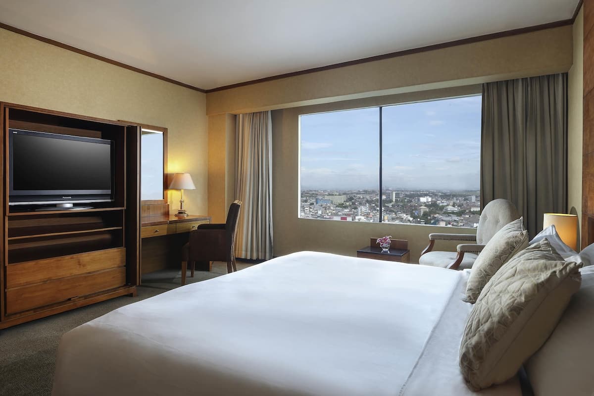 Une chambre d’hôtel avec un grand lit, un bureau avec une télévision, une chaise, une fenêtre donnant sur la ville et une table de chevet avec une lampe.