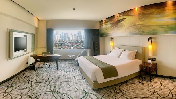 A hotel room with a large bed, a desk, a chair, a TV, and a view of a cityscape. - Grand Mercure Medan Angkasa (Medan)