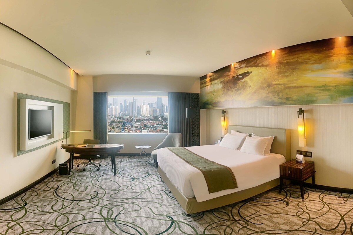 Habitación de hotel con una cama grande, un escritorio, una silla, un televisor y vistas a la ciudad.
