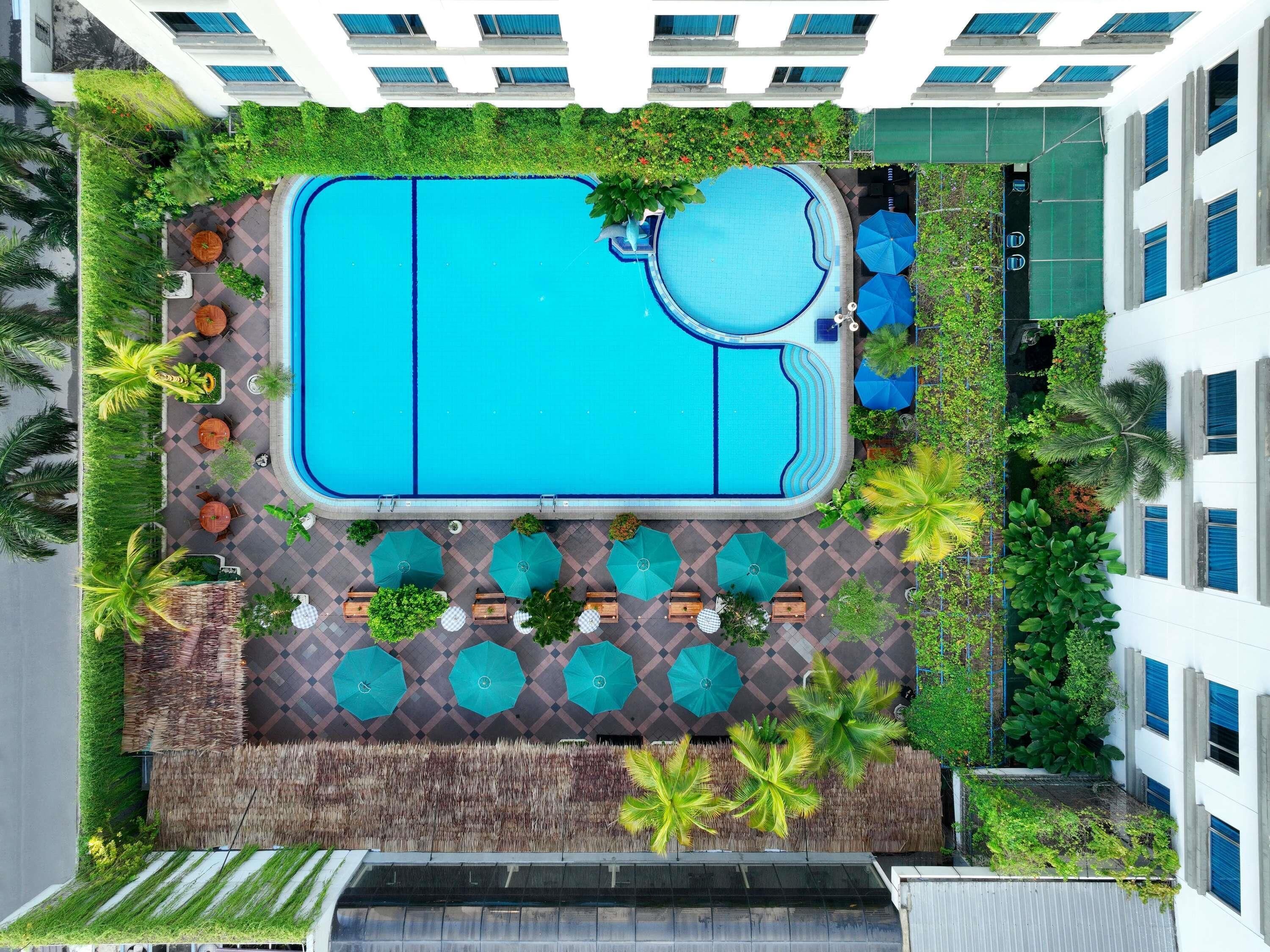 An aerial view of a hotel with a large rectangular pool, surrounded by palm trees and a patio area with umbrellas and seating.