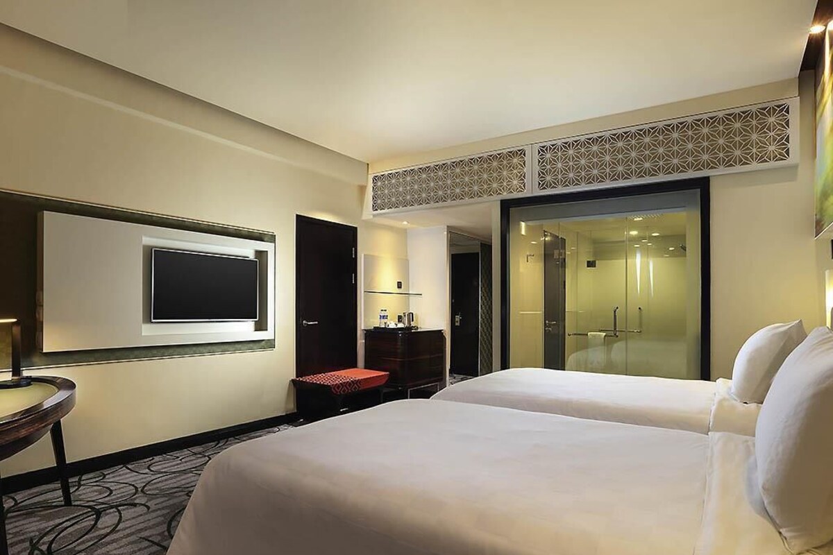 Habitación de hotel con dos camas, televisión, escritorio, silla, ducha y puerta de vidrio.