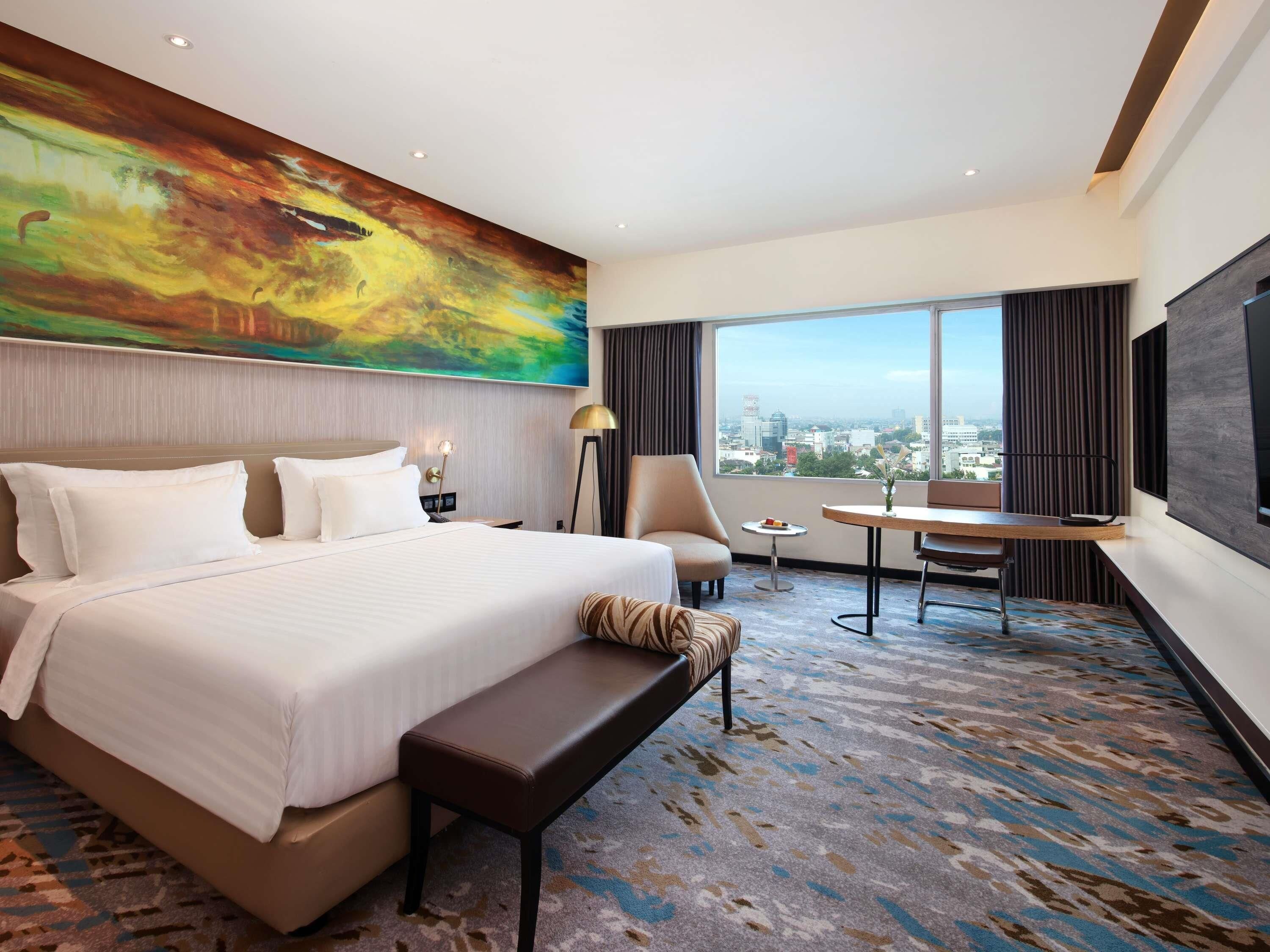 A hotel room with a large bed, a desk, a chair, a bench, a painting on the wall, and a view of the city through large windows.