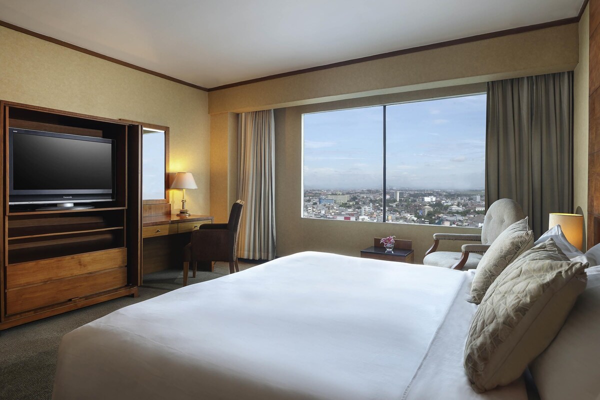 A hotel room with a large bed, a desk with a television, a chair, a window with a city view, and a nightstand with a lamp.