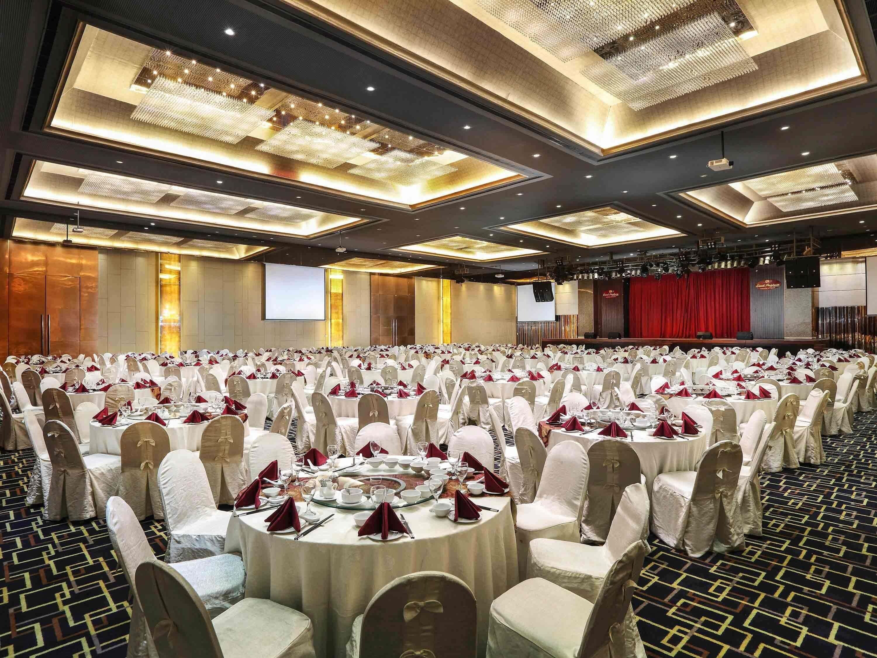 Un amplio salón de banquetes con mesas redondas, sillas blancas y un escenario con cortina roja.