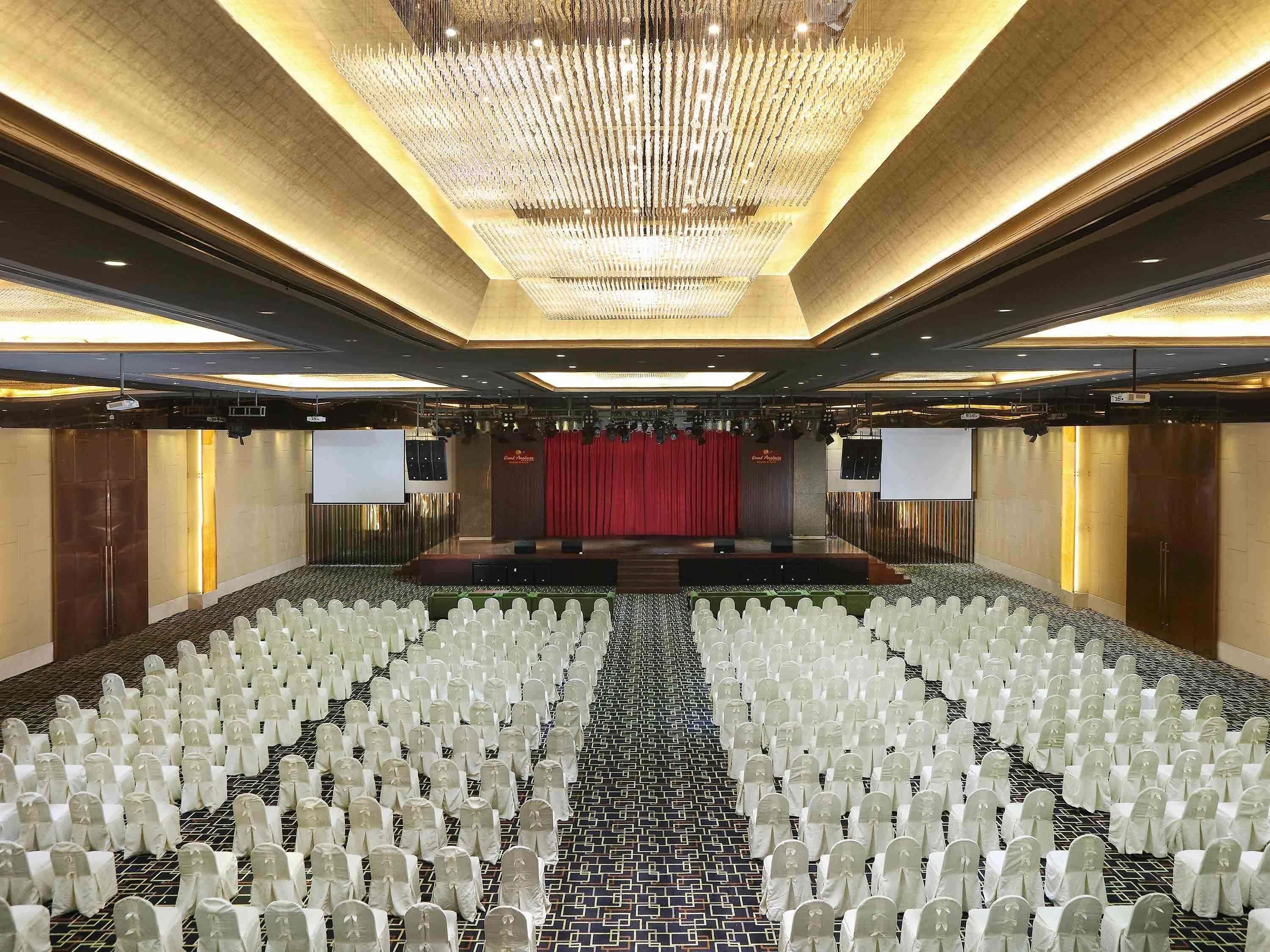 A large conference room with rows of chairs, a stage with a red curtain, and a chandelier.