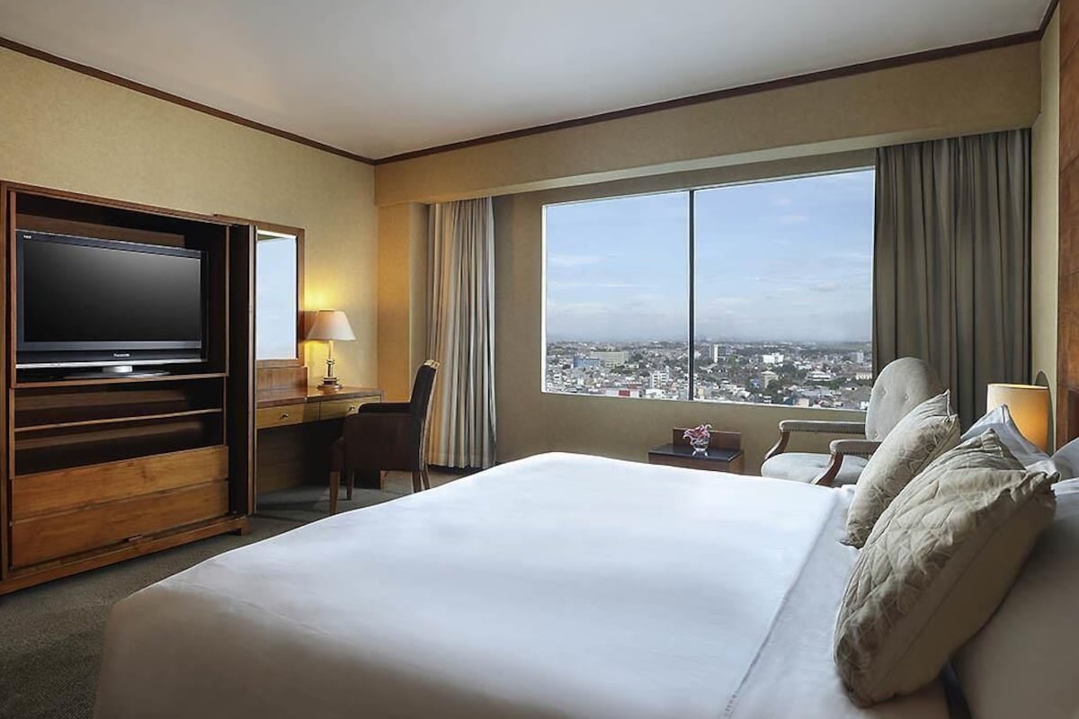 Habitación de hotel con una cama grande, televisor, escritorio, silla, lámpara y vista a la ciudad a través de un ventanal.