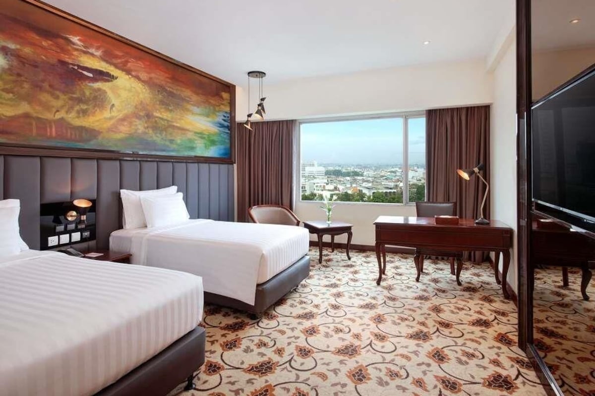 Habitación de hotel con dos camas, un escritorio, un televisor, un ventanal amplio con vistas a la ciudad y un cuadro en la pared.