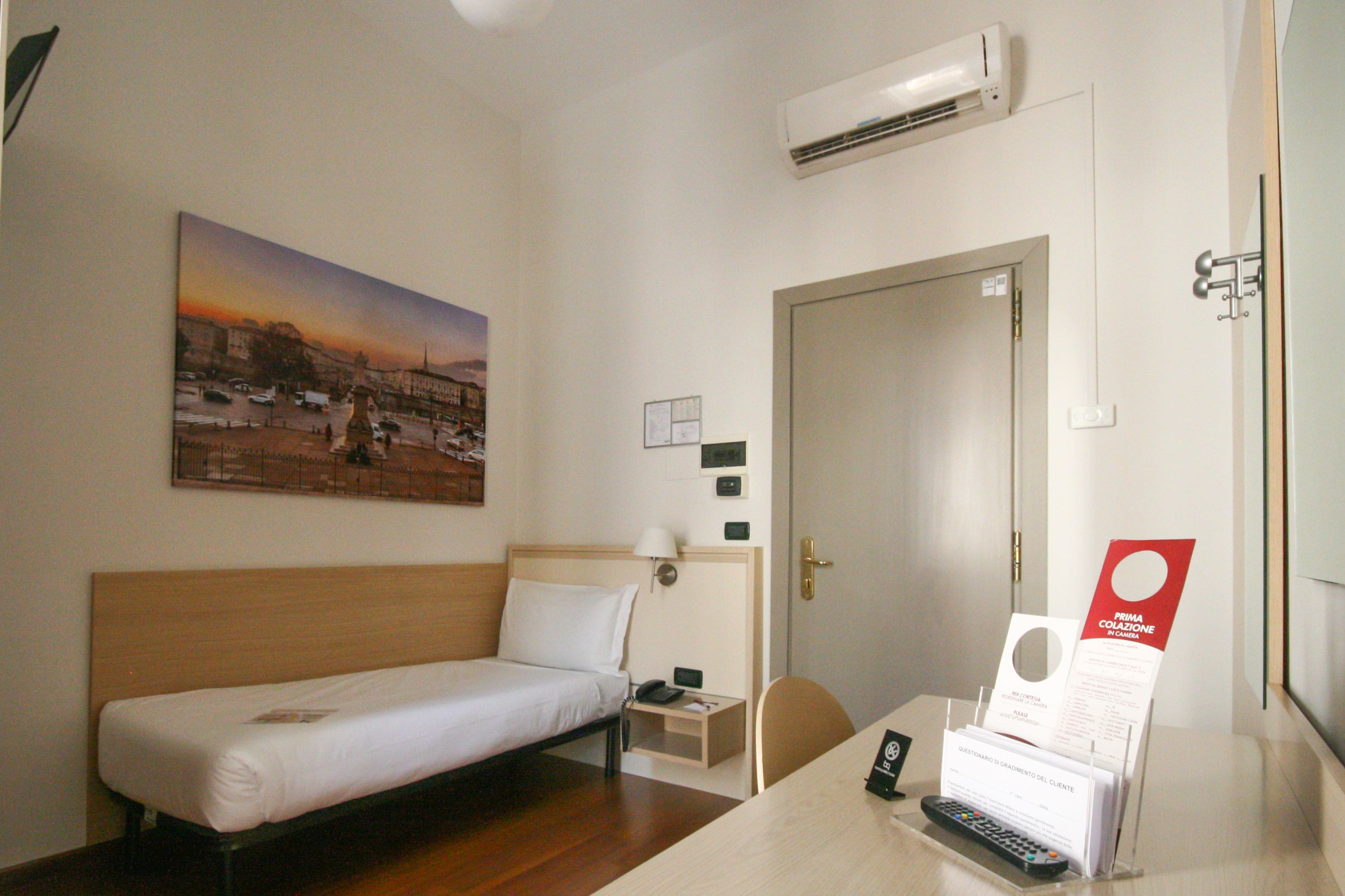 Une chambre d’hôtel avec un lit simple, un bureau avec une chaise, une table de chevet, un climatiseur fixé au mur et un tableau représentant un coucher de soleil.