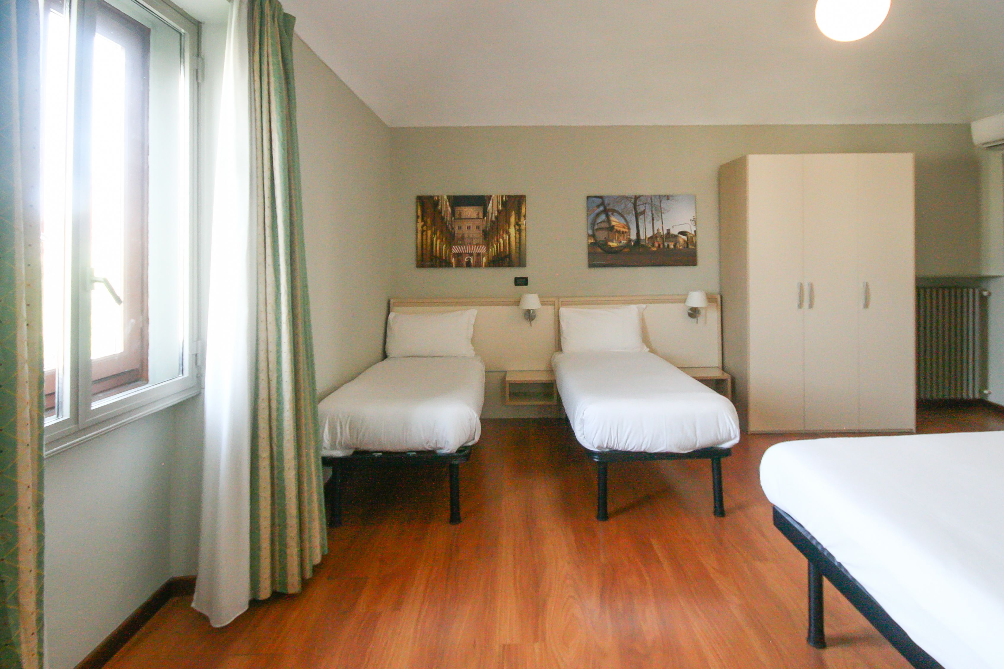 Ein Hotelzimmer mit zwei Einzelbetten, einem großen Kleiderschrank, einem Fenster mit Vorhängen und zwei Wandleuchten.