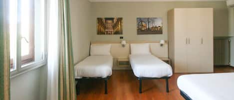 A hotel room with two single beds, a large wardrobe, a window with curtains, and two wall-mounted lamps.
