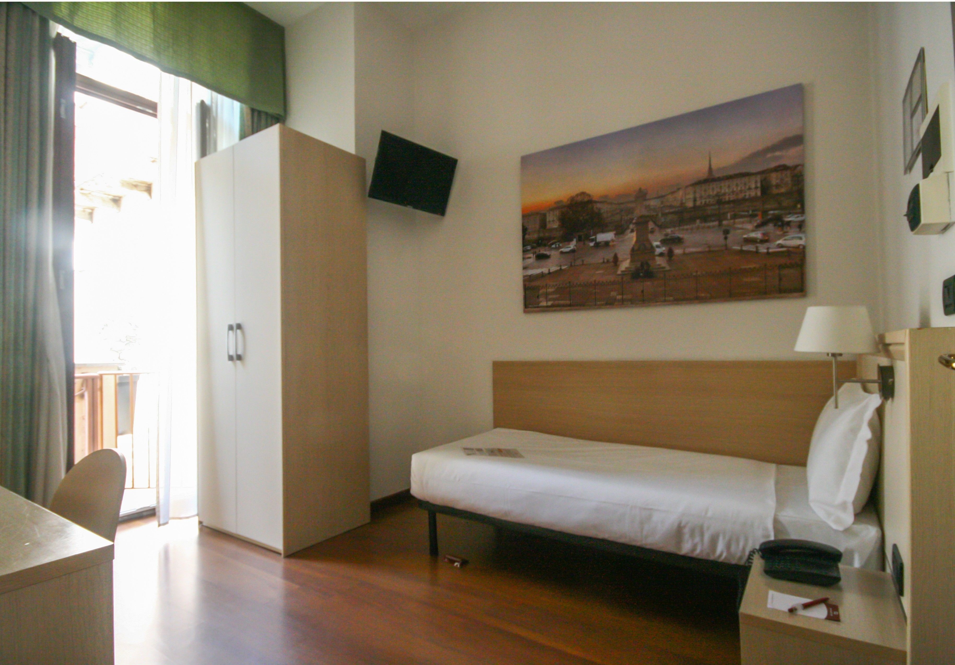 A hotel room with a single bed, a desk, a chair, a wardrobe, a flat-screen TV mounted on the wall, and a painting depicting a sunset scene.