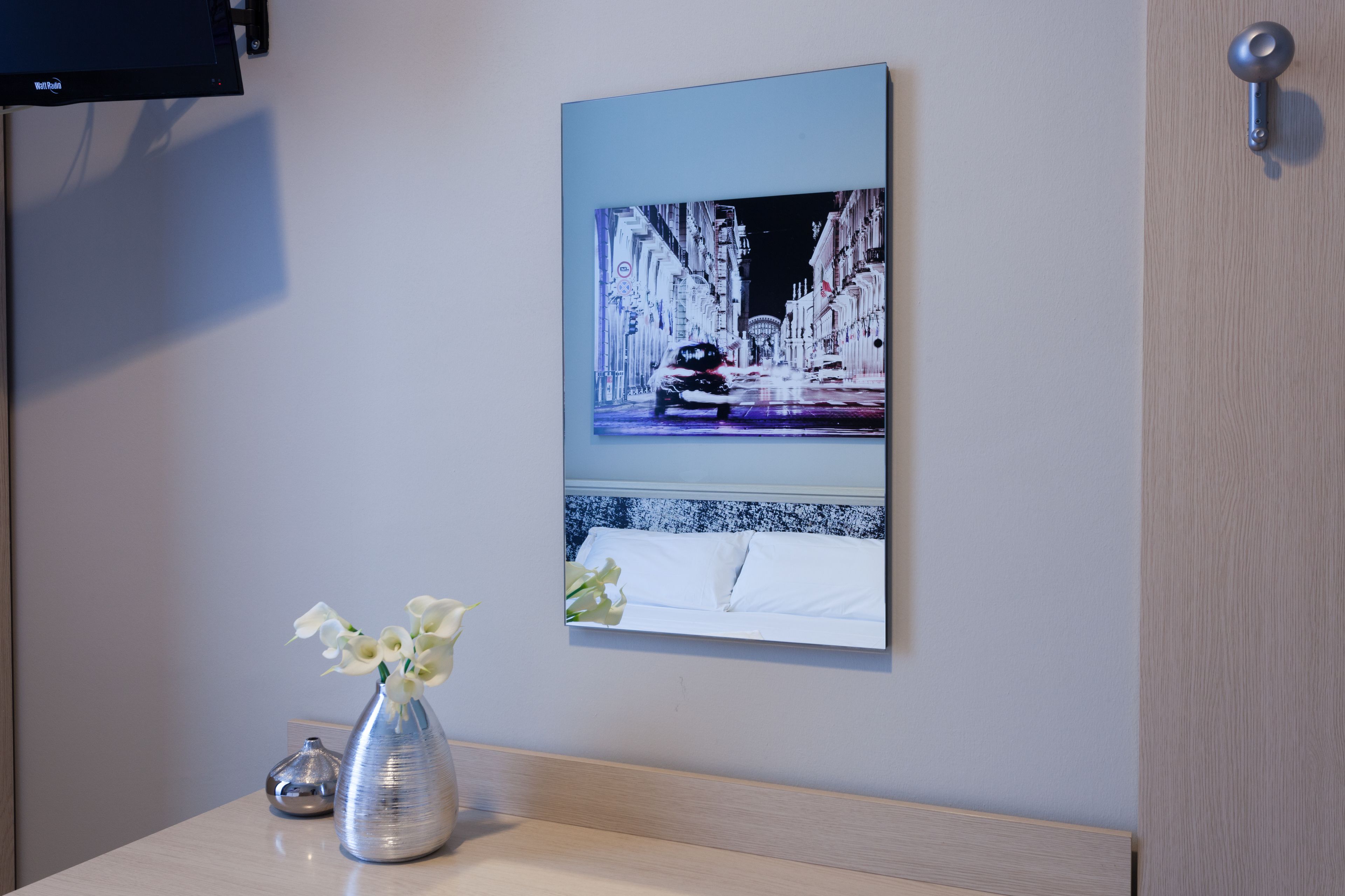 A hotel room with a bed, a vase with flowers, a painting of a snowy street scene, and a television mounted on the wall.