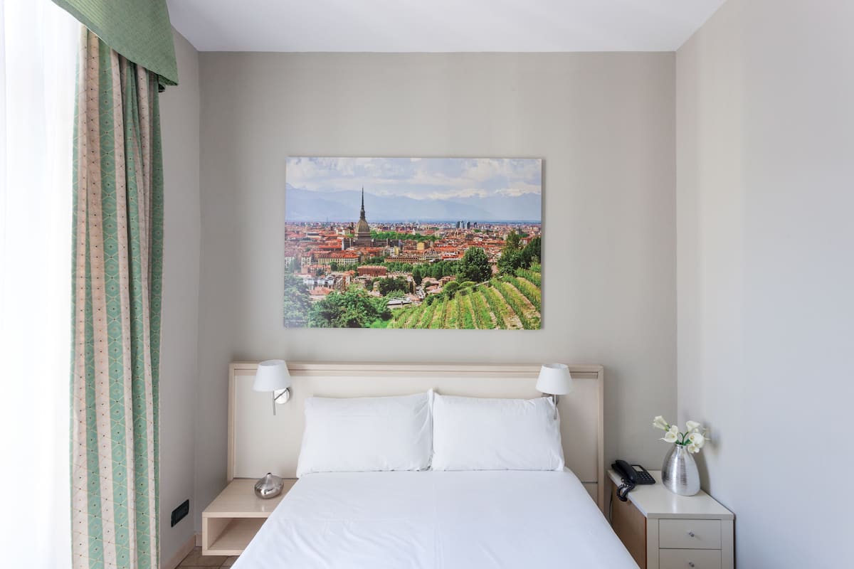 A hotel room with a large bed, a bedside table with a lamp, a nightstand with a phone, and a painting of a cityscape on the wall.