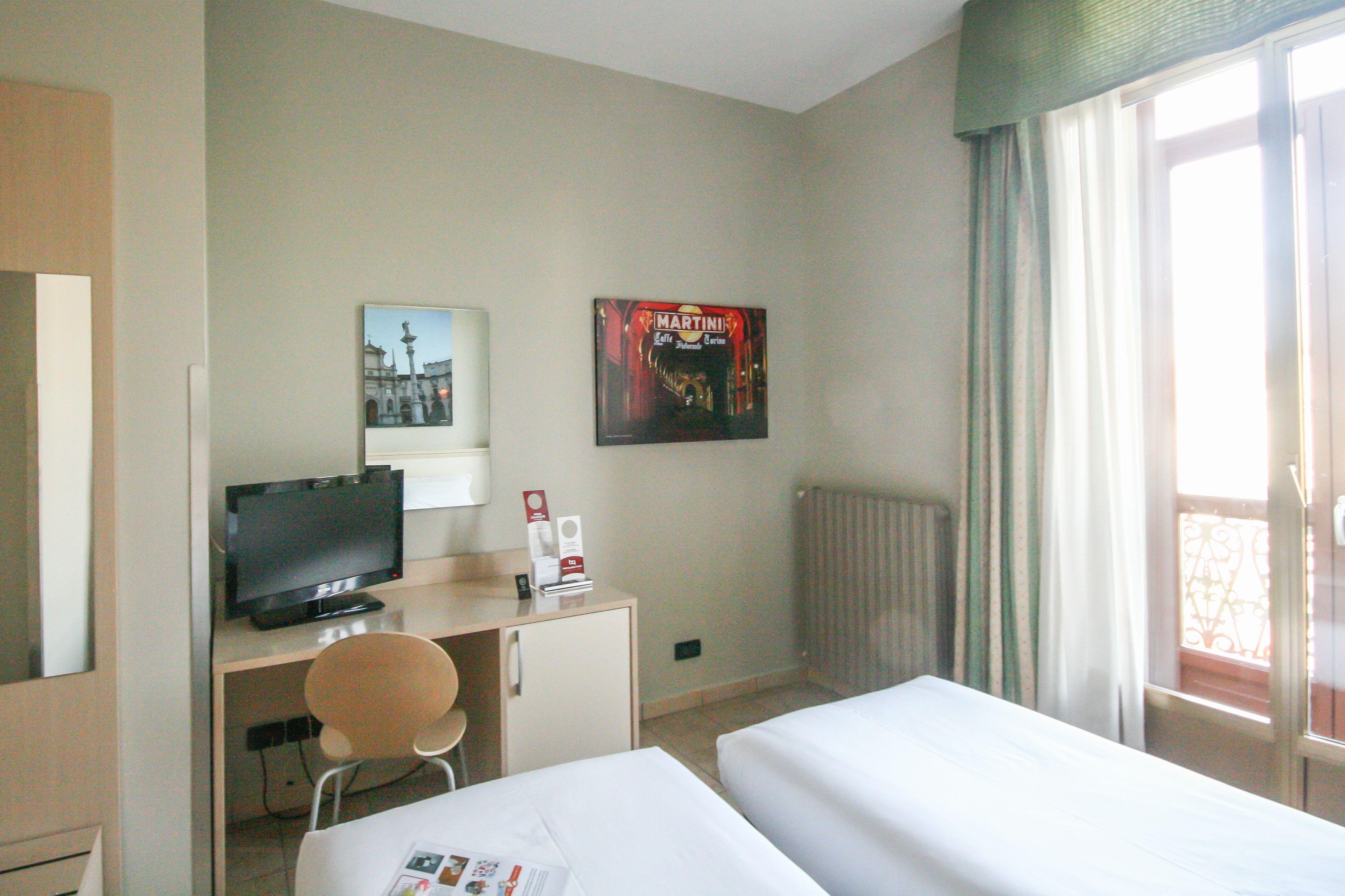 Habitación de hotel con cama, escritorio con televisor, silla, radiador y ventana con cortinas.