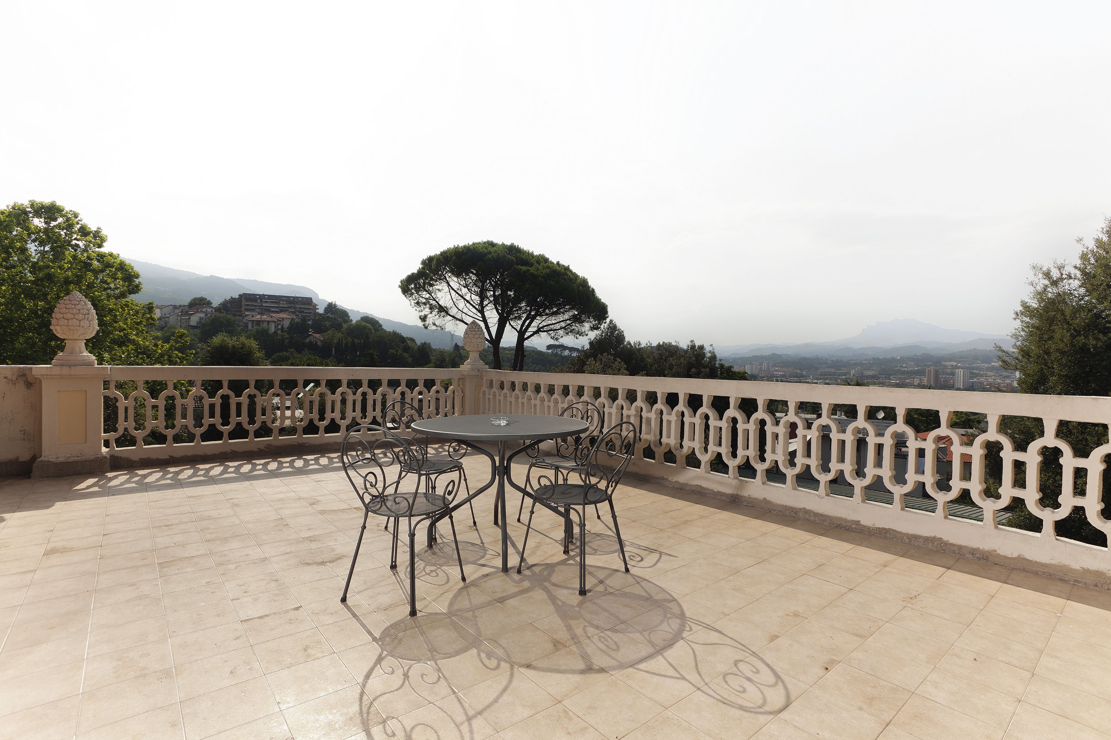 Una terraza con una mesa y sillas, una barandilla decorativa y vistas a un árbol y edificios a lo lejos.