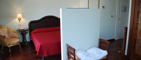 A hotel room with a bed, a wooden chair, a small table with a lamp, and a door.
