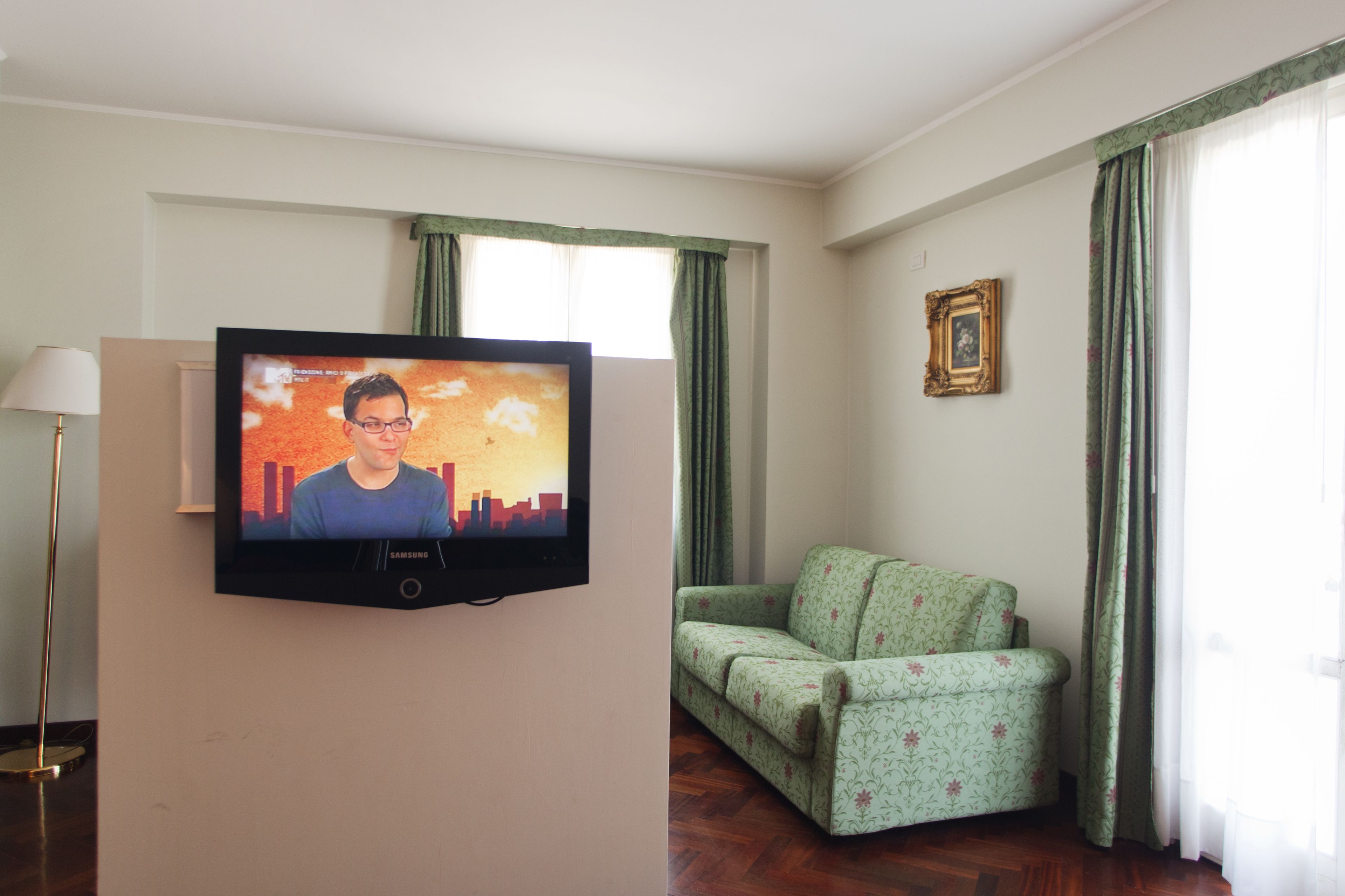 A room with a television mounted on a white wall, a green patterned sofa, a floor lamp, and a framed picture on the wall.