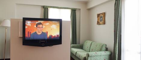 A room with a television mounted on a white wall, a green patterned sofa, a floor lamp, and a framed picture on the wall.