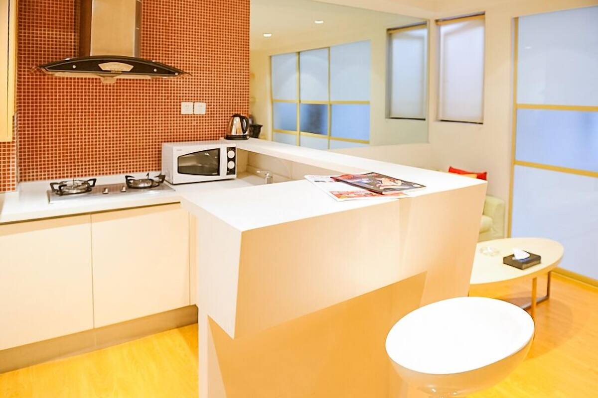 A modern kitchen with a white countertop, a gas stove, a microwave, and a kettle. There is a small island with a white sink and a round table with a chair.