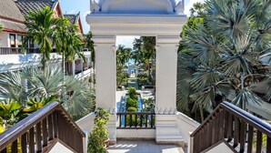 A view through a white archway with columns, leading to a pool area surrounded by palm trees and a building with a tiled roof. - Beyond Samui (Koh Samui)