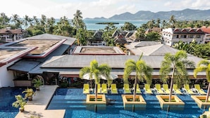 A resort pool with clear blue water, surrounded by lounge chairs and palm trees, overlooking a coastal area with buildings and mountains in the background. - Beyond Samui (Koh Samui)