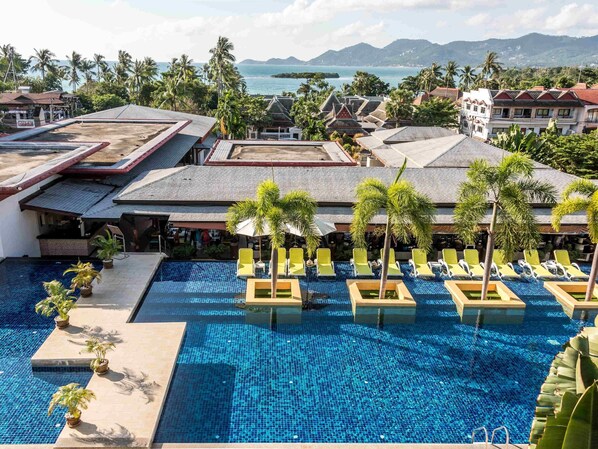 A resort pool with clear blue water, surrounded by lounge chairs and palm trees, overlooking a coastal area with buildings and mountains in the background. - Beyond Samui (Koh Samui)