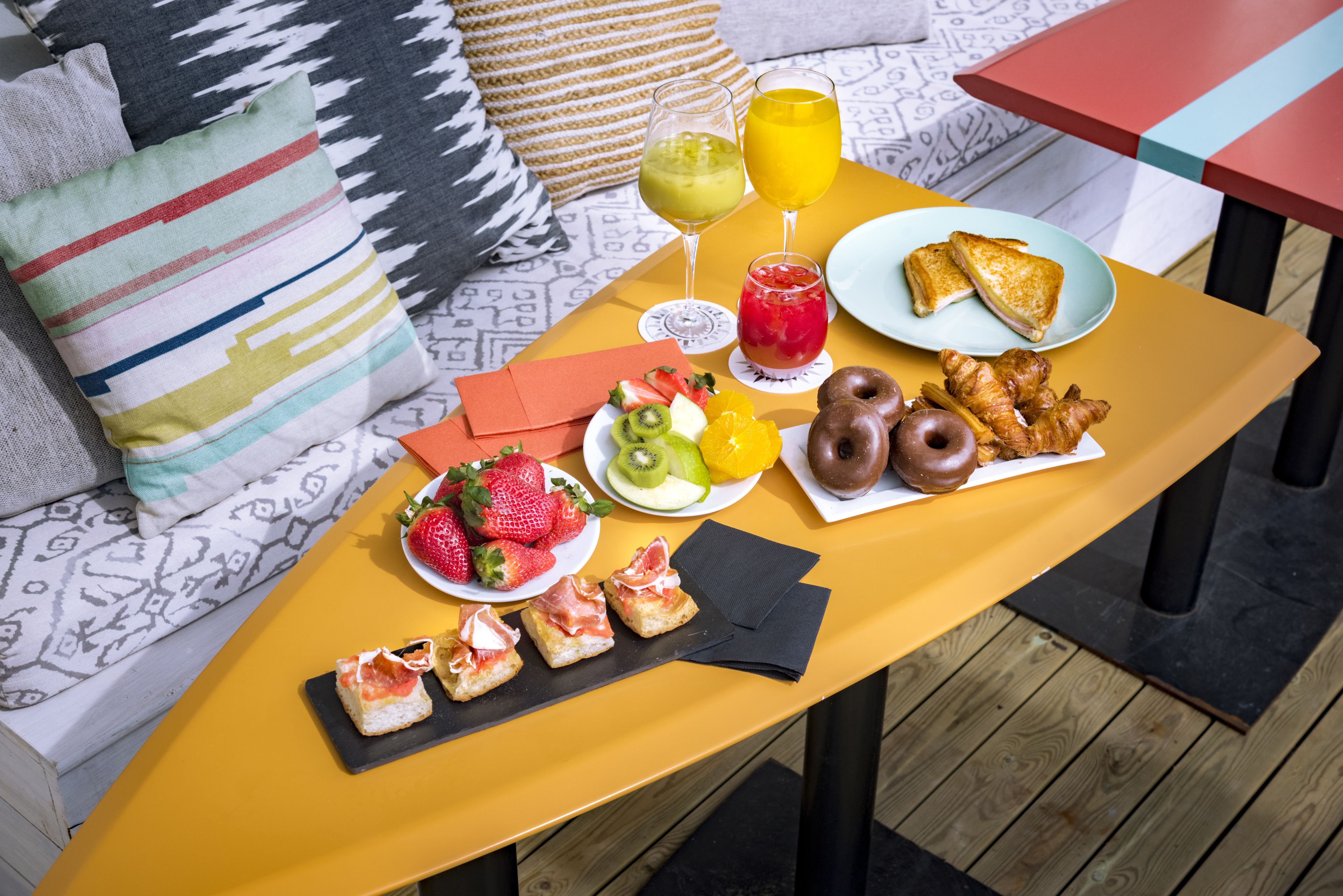 Una mesa de comedor con comida y bebidas, un sofá con cojines estampados y una terraza de madera.
