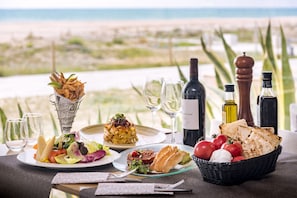 Una mesa dispuesta con platos de comida, vasos y botellas de vino, con vista a una playa.