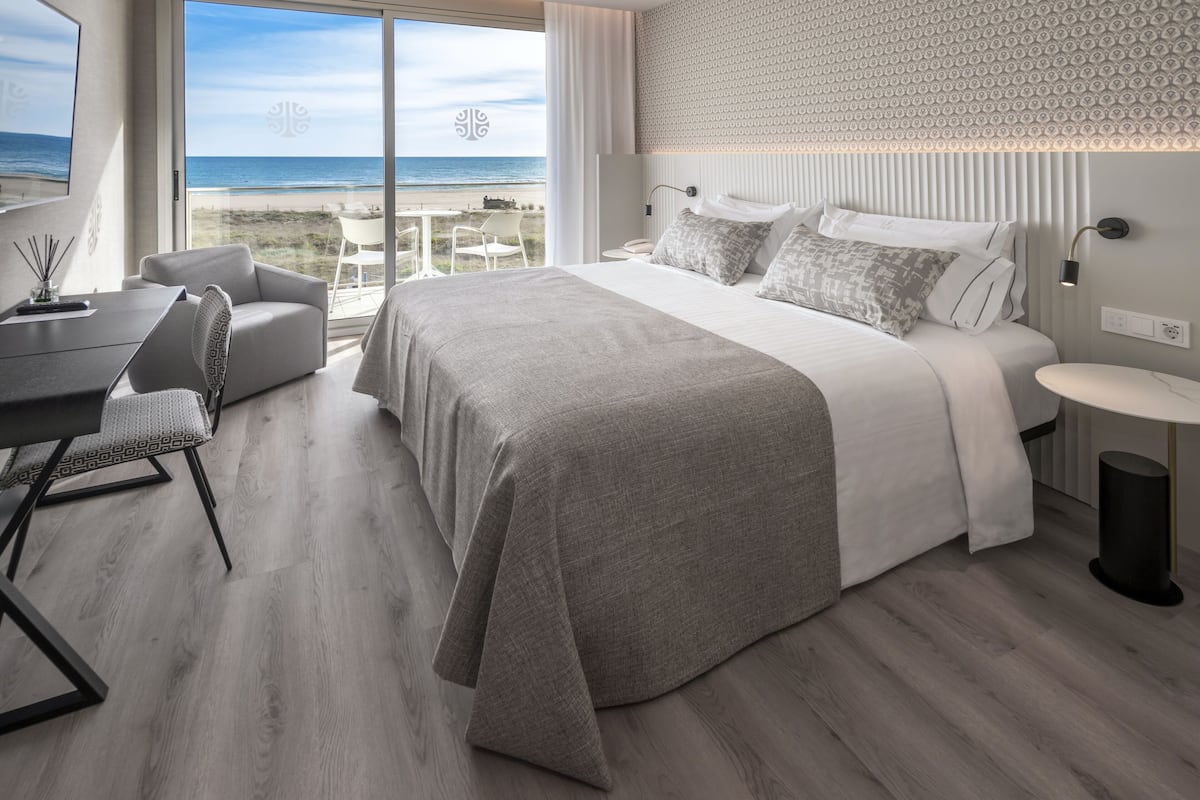 A modern bedroom with a large bed, a desk, and a view of the beach.
