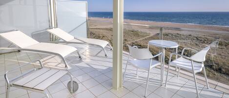 A balcony with a view of the beach, white lounge chairs, and a small table.