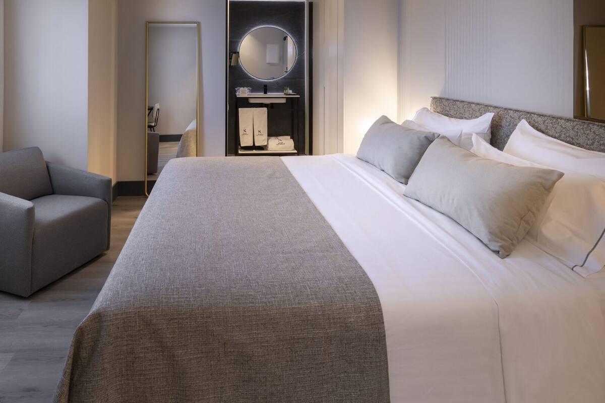 A hotel room with a large bed, a chair, a mirror, and a small table with toiletries.