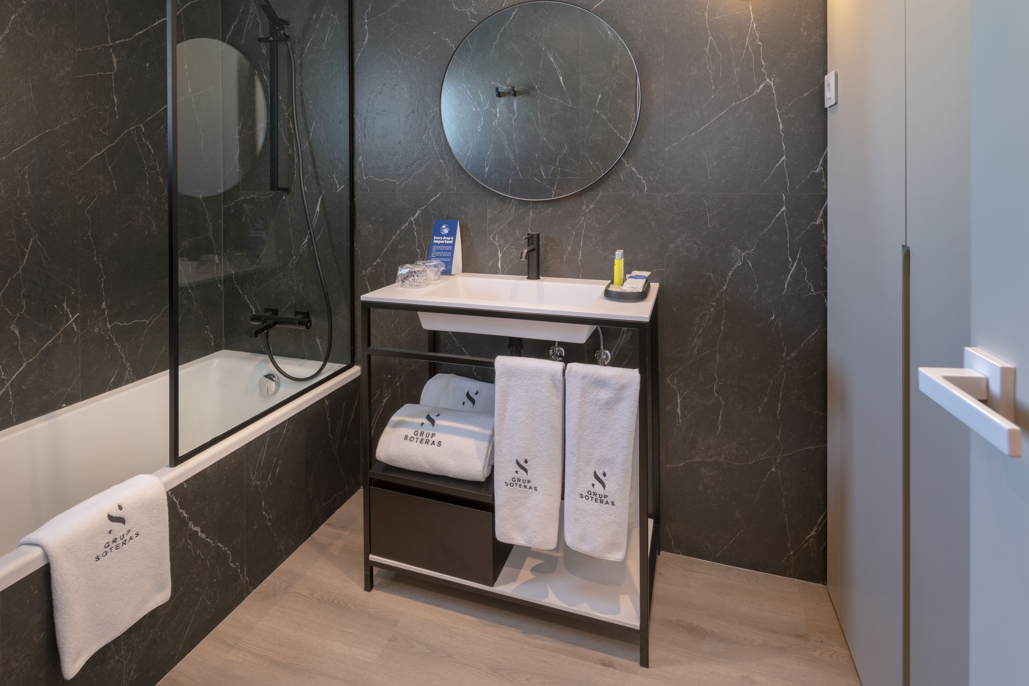 Un baño moderno con pared de mármol, espejo redondo, lavamanos blanco, cabina de ducha con mampara de vidrio y toallero con toallas etiquetadas como "GRUP SOTERAS".