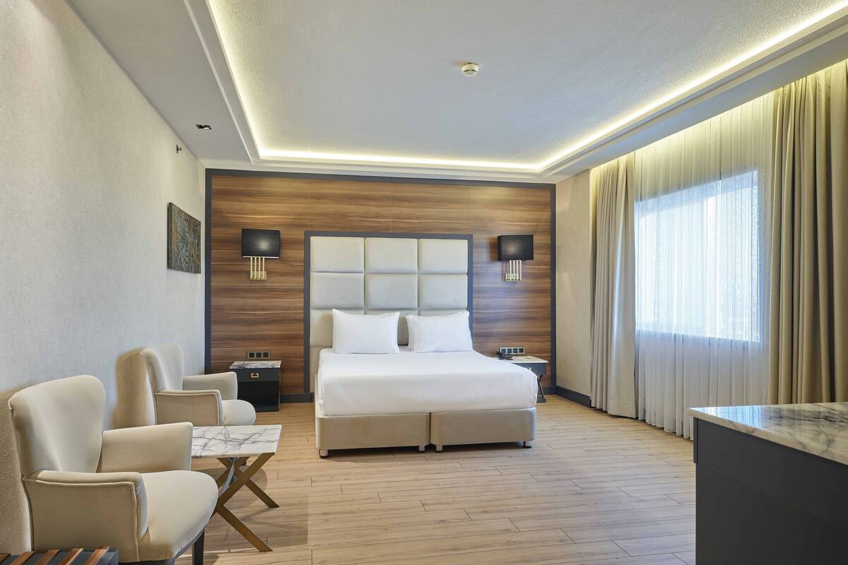 Moderni hotellihuone, jossa on suuri sänky, kaksi yöpöytää, työpöytä, tuoli ja suuri ikkuna, jossa on verhot.
