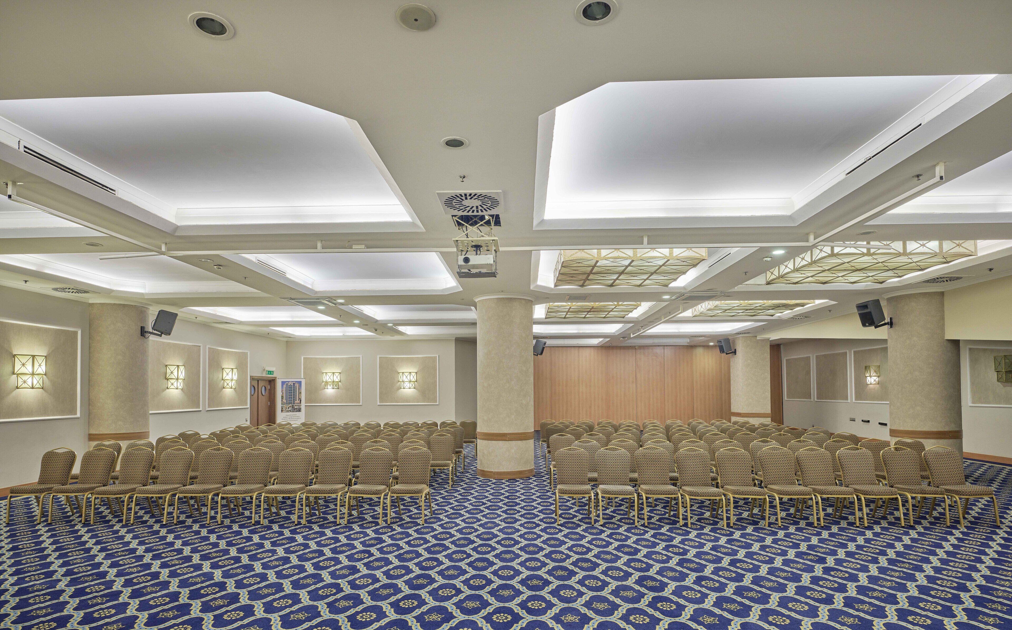 Un amplio salón de conferencias con filas de sillas, una alfombra estampada y un techo alto con tragaluces.