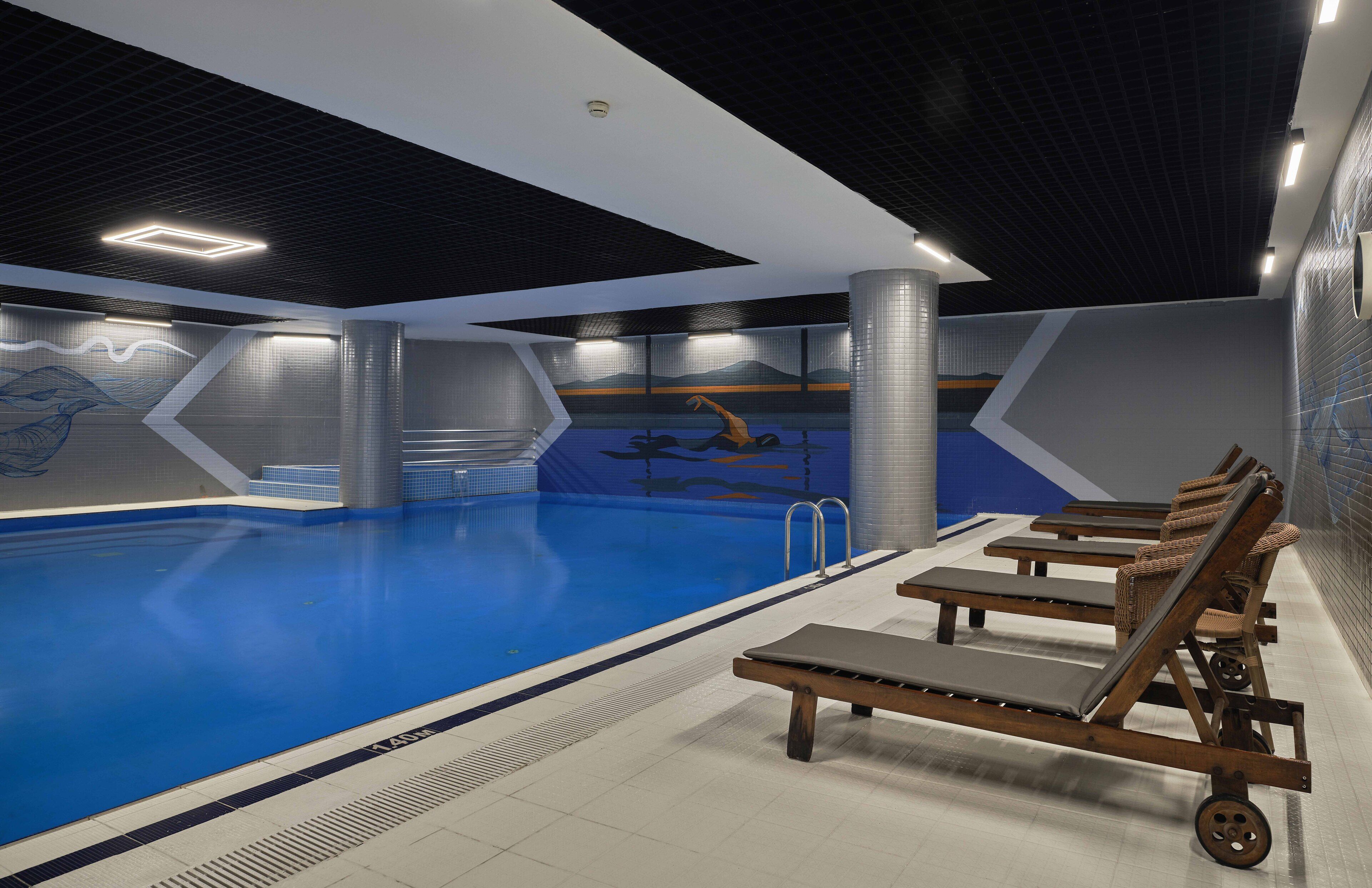 An indoor swimming pool with clear lane markings, a mural on the wall, and lounge chairs along the poolside.