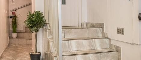 Un pasillo moderno y limpio con piso de mármol, una planta en maceta y una escalera que lleva a una puerta. Un tapete verde con la inscripción “Little Palace” está colocado al pie de la escalera.