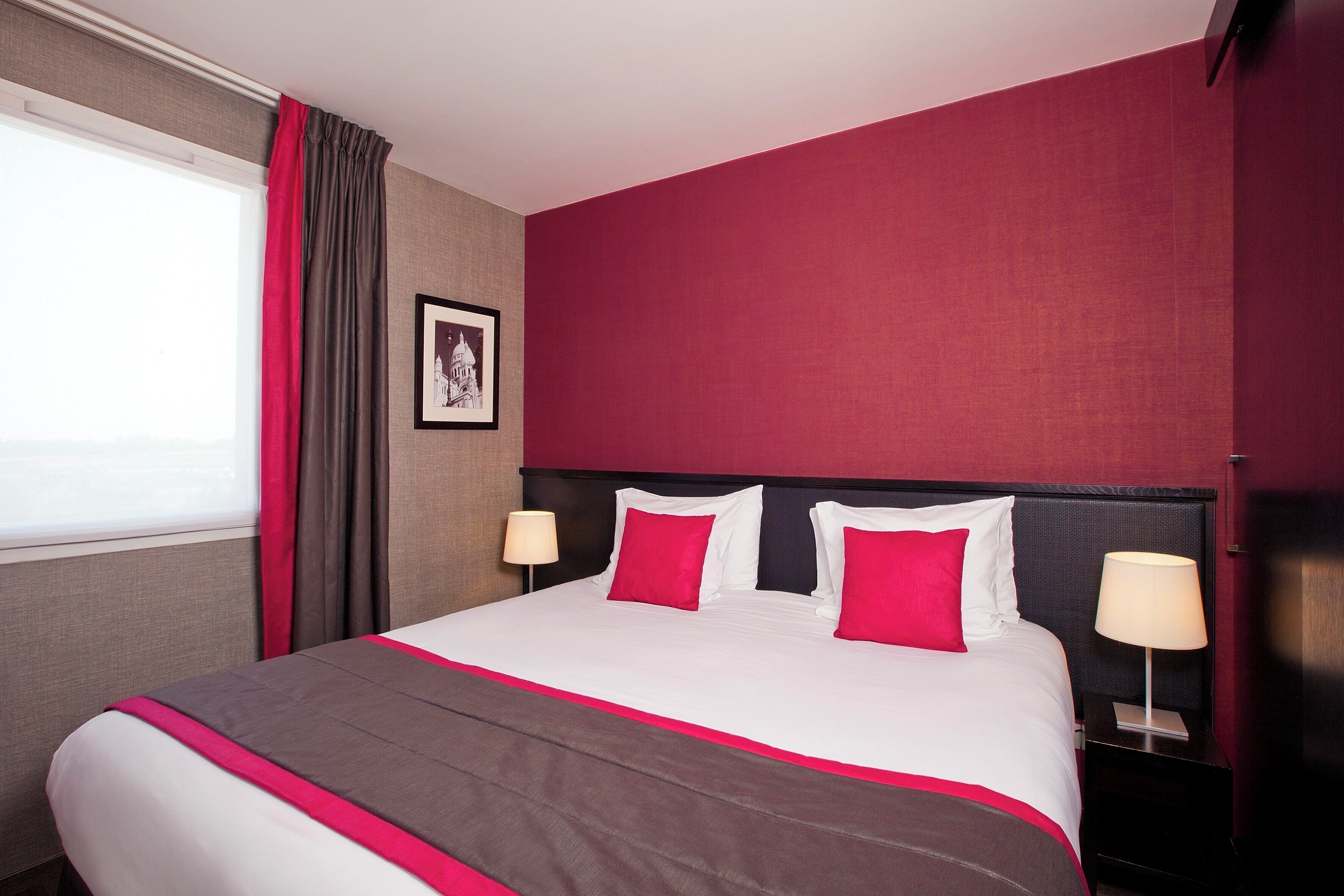 A hotel room with a large bed, red and white bedding, a nightstand with a lamp, and a framed picture on the wall.
