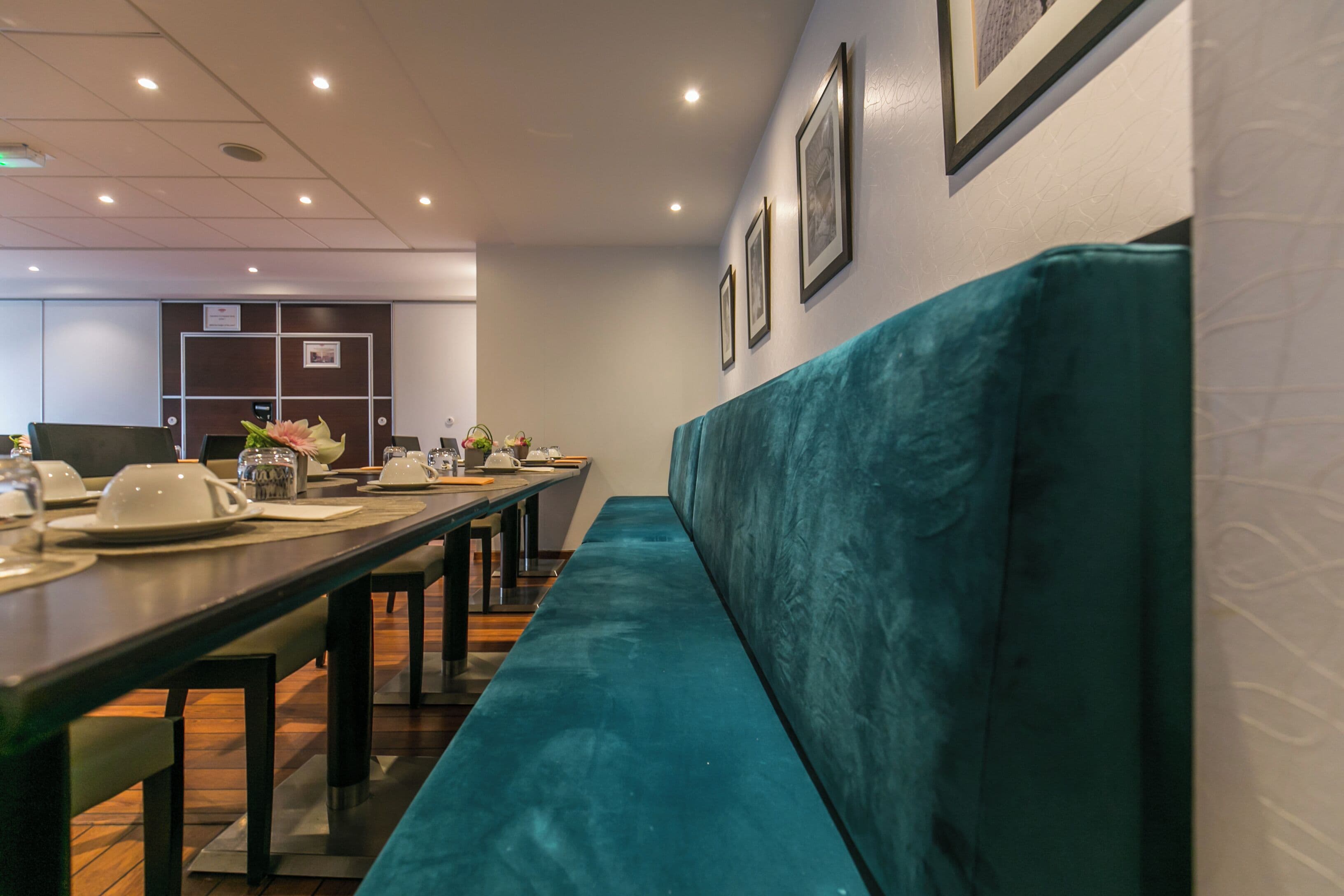 A restaurant with a long table, teal cushioned bench, and framed pictures on the wall.