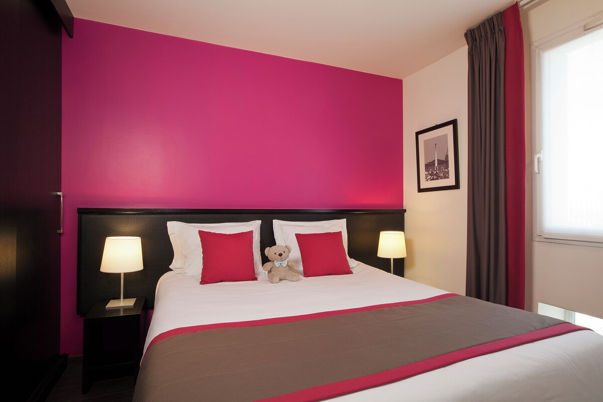 Une chambre d’hôtel avec un grand lit, une literie rouge et blanche, deux lampes de chevet et un ours en peluche sur le lit.