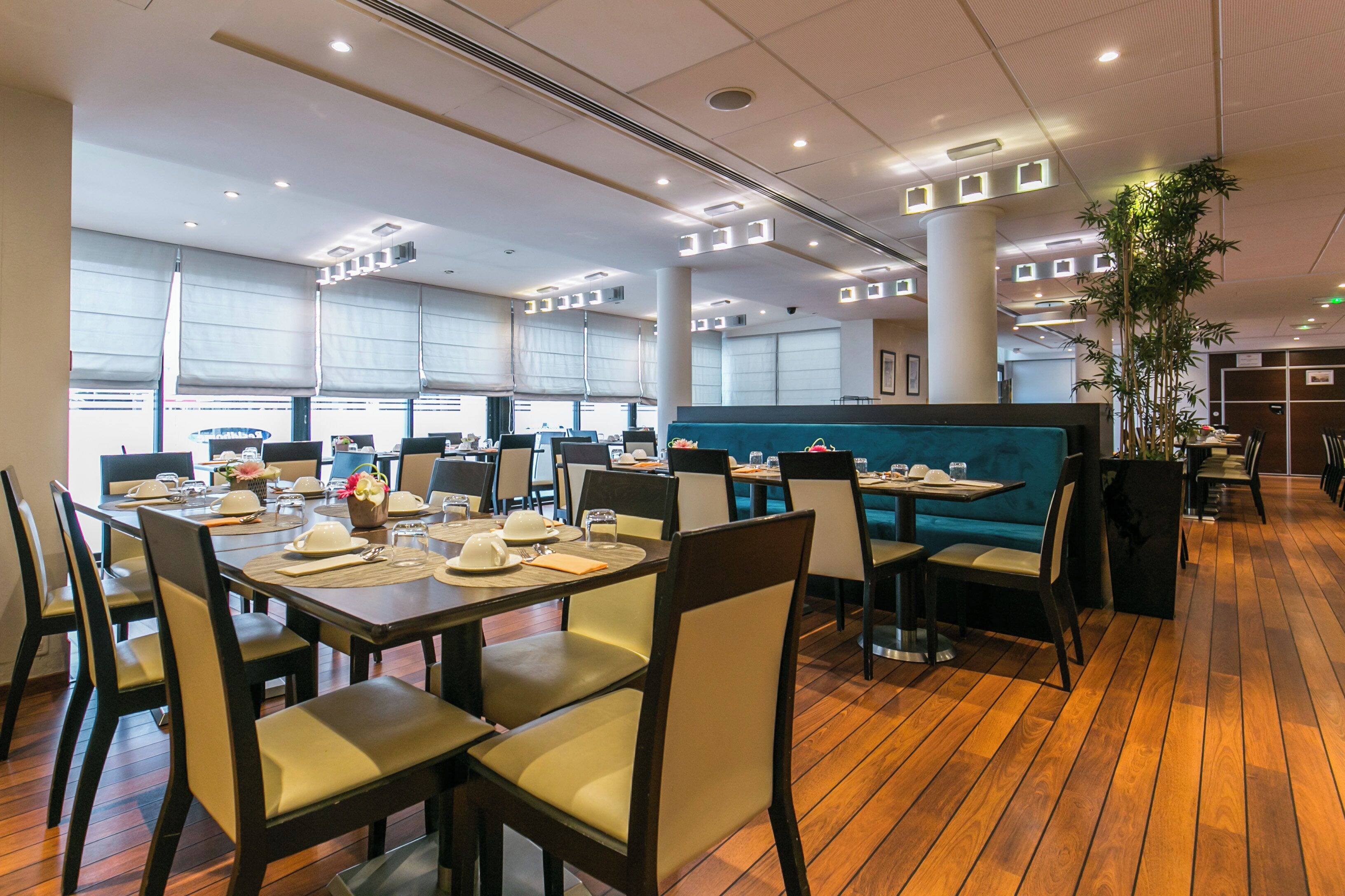 A spacious restaurant with wooden flooring, multiple dining tables with chairs, and large windows allowing natural light.
