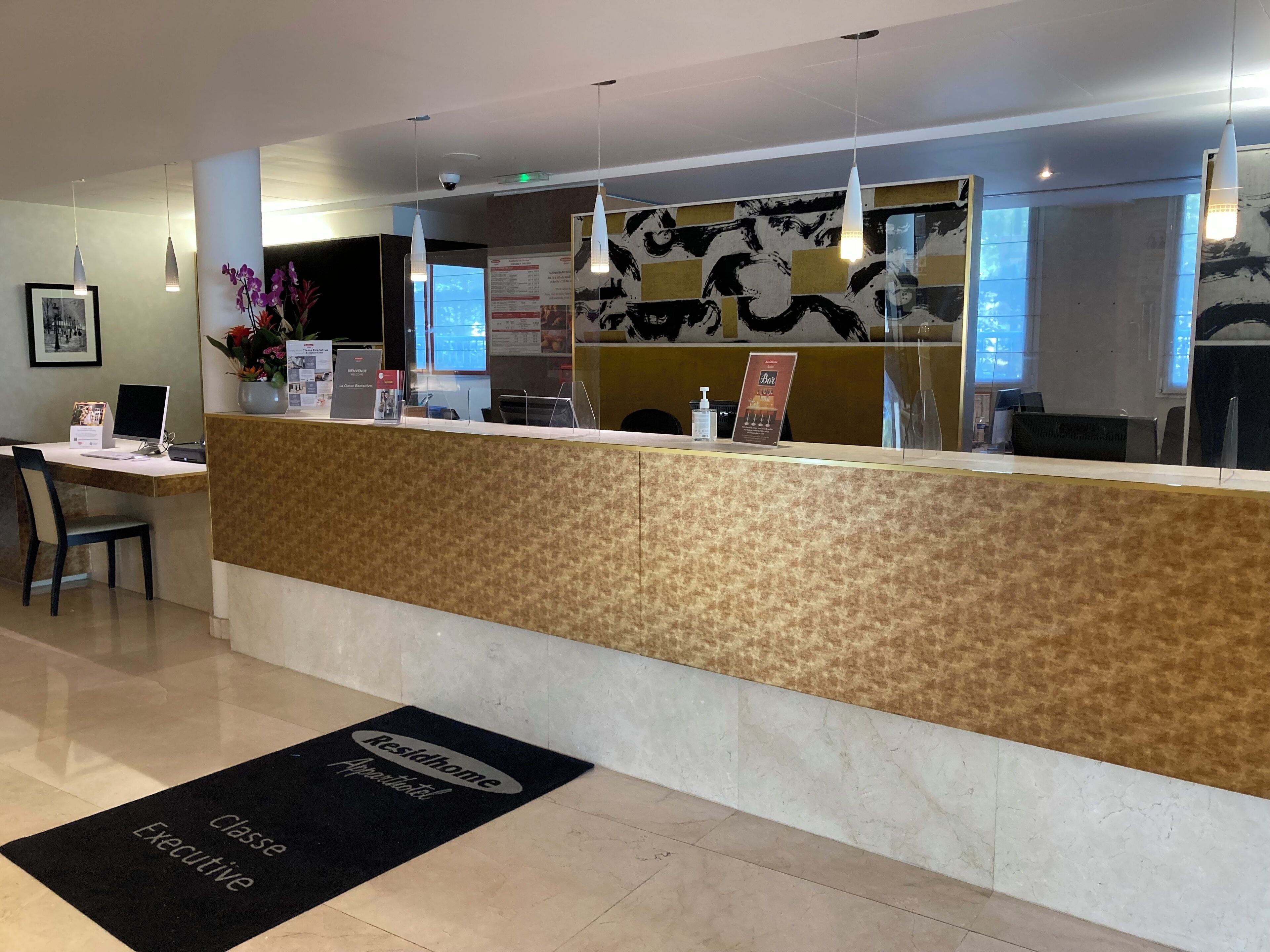 Un espace d’accueil d’hôtel comprenant un comptoir de réception, un ordinateur, une chaise et un paillasson noir indiquant l’entrée du salon VIP.
