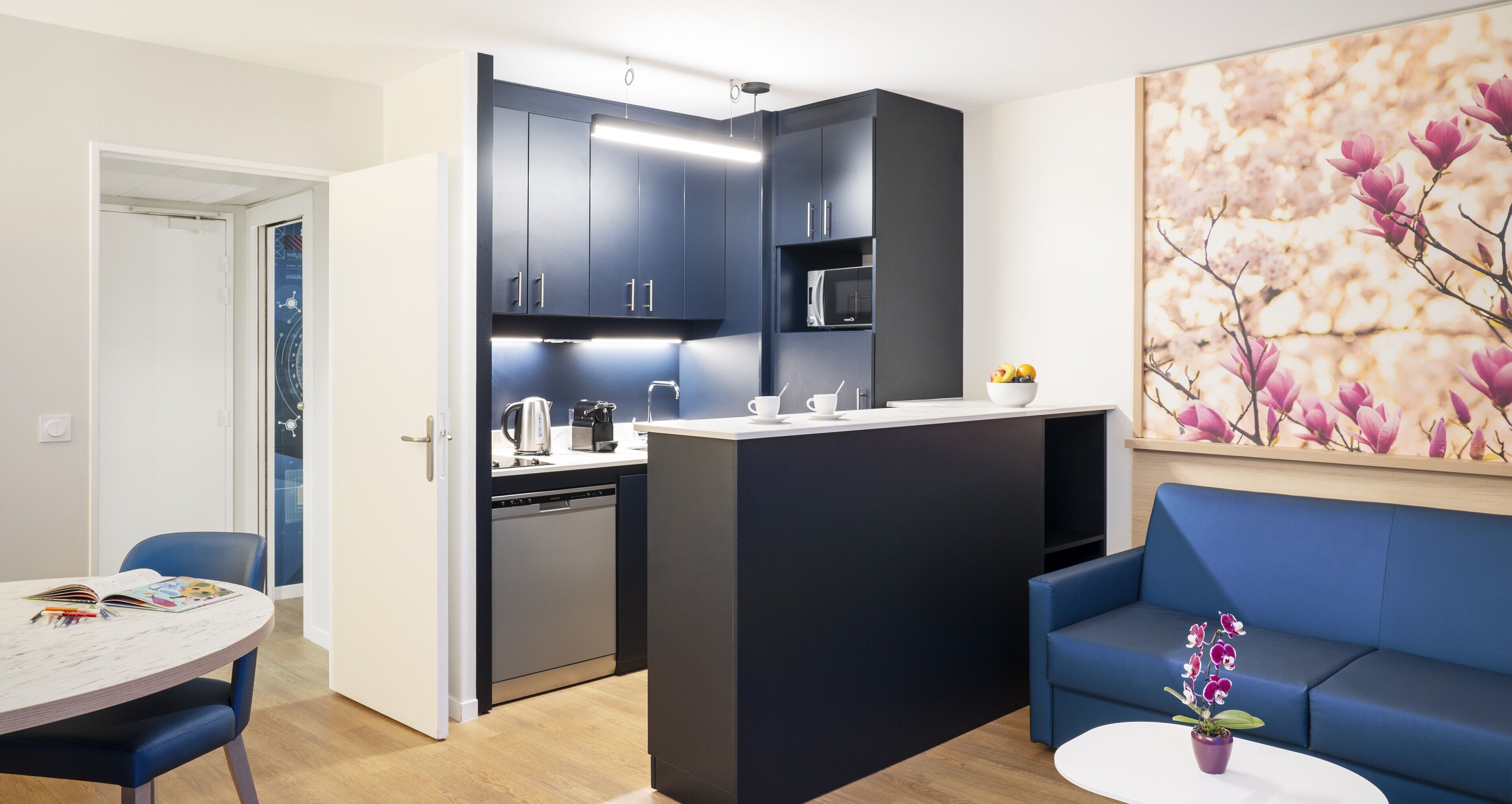 A modern kitchen with dark cabinets, a dishwasher, and a microwave. There is a dining area with a table and chairs, and a blue sofa.