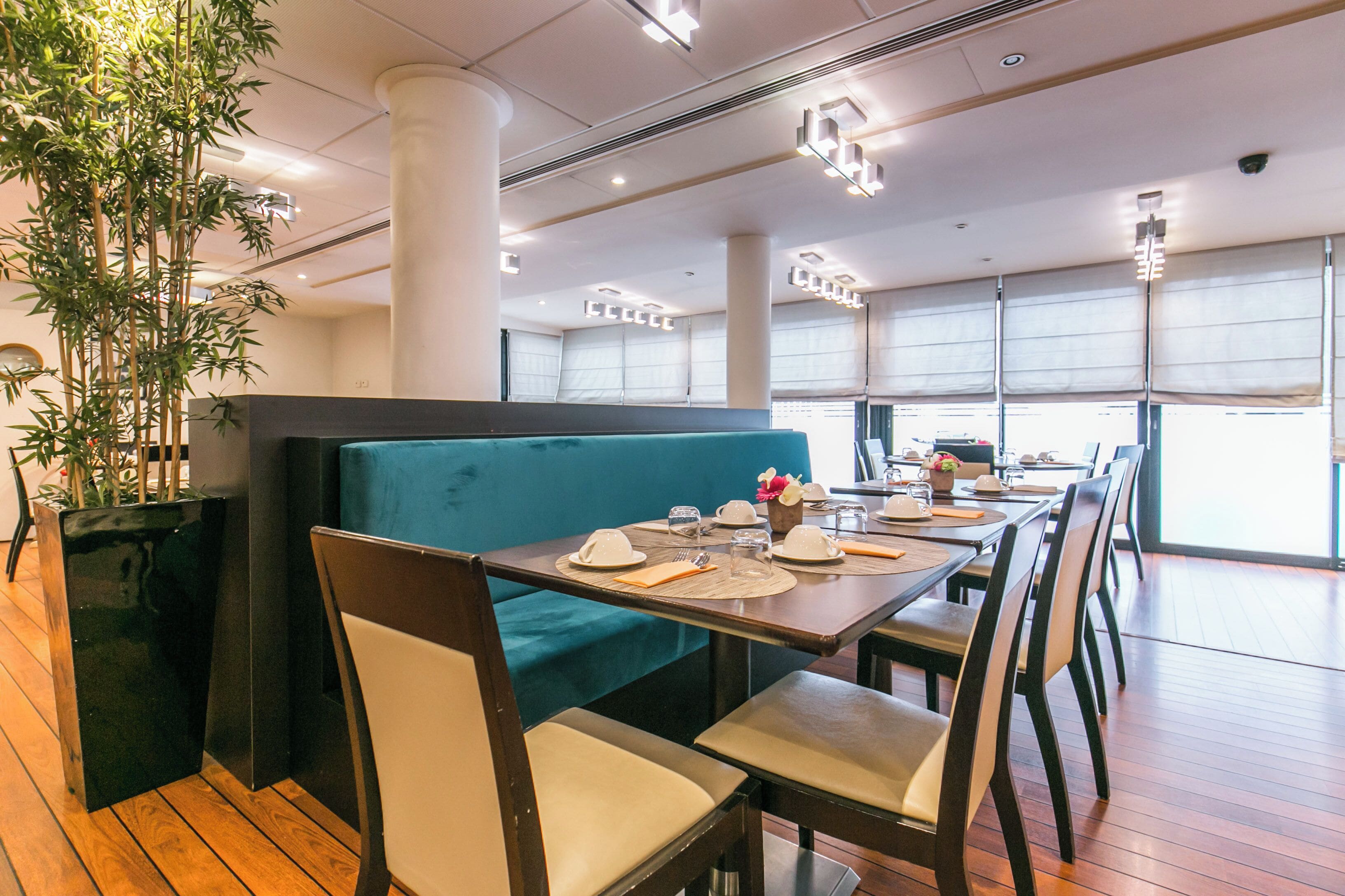 A restaurant with a long wooden table set for dining, a green booth seating area, and a large potted plant.