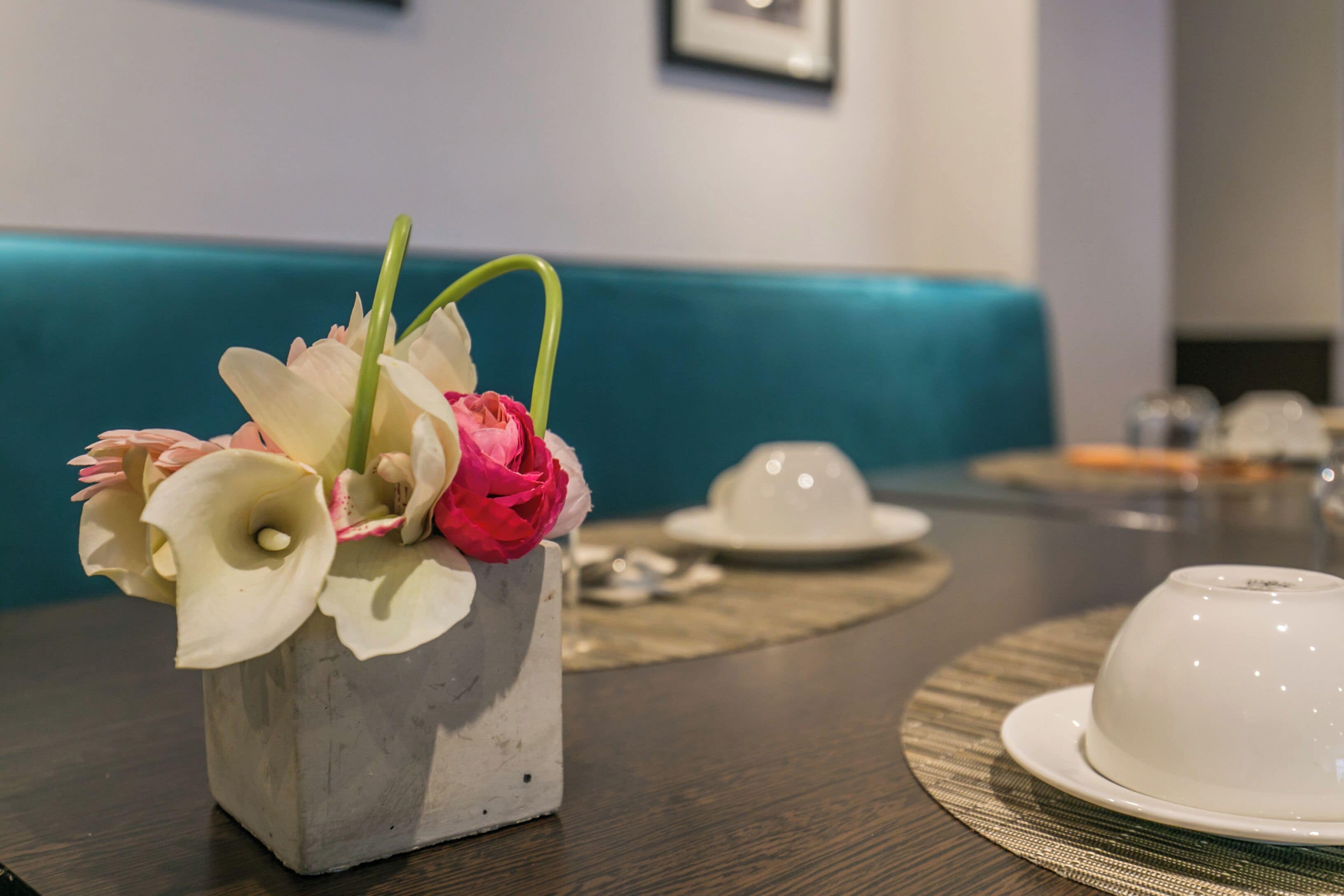 A table with a white teacup and saucer, a white vase with flowers, and a teal-colored bench in the background.