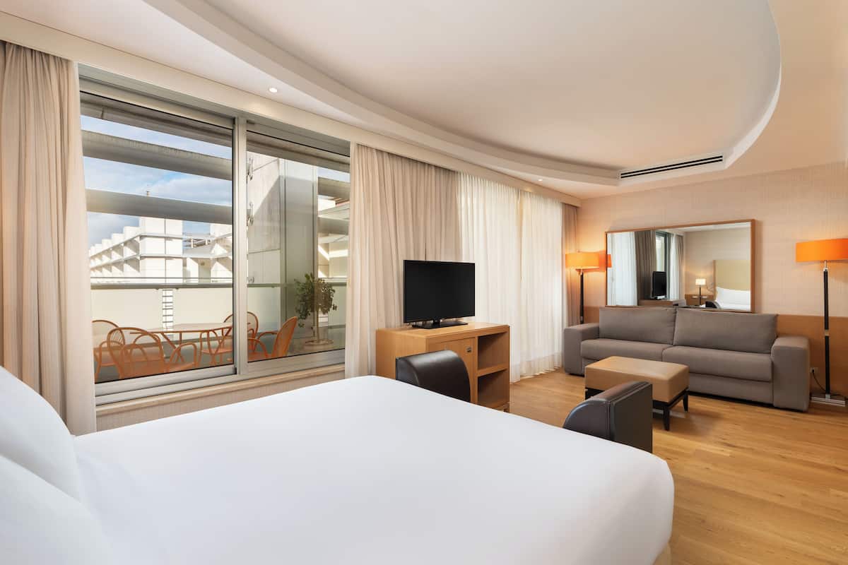 A hotel room with a large bed, a desk with a TV, a sofa, and a balcony with orange chairs.