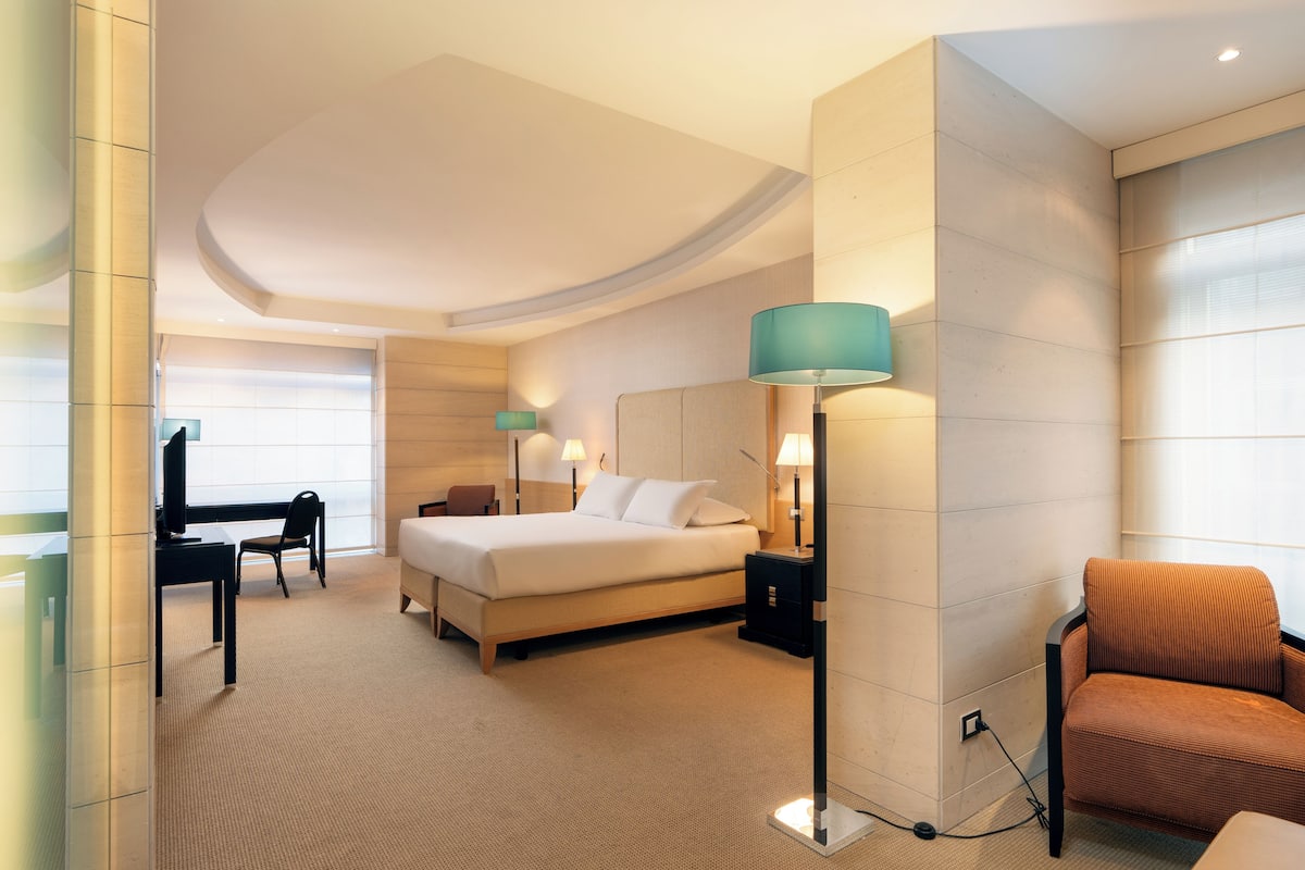 A hotel room with a bed, desk, chair, lamp, and a view of the outside.