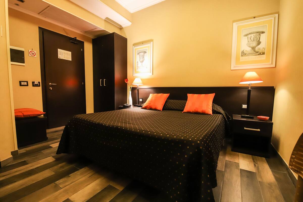 Una habitación de hotel con una cama, mesitas de noche, una puerta y una pared decorada con cuadros.
