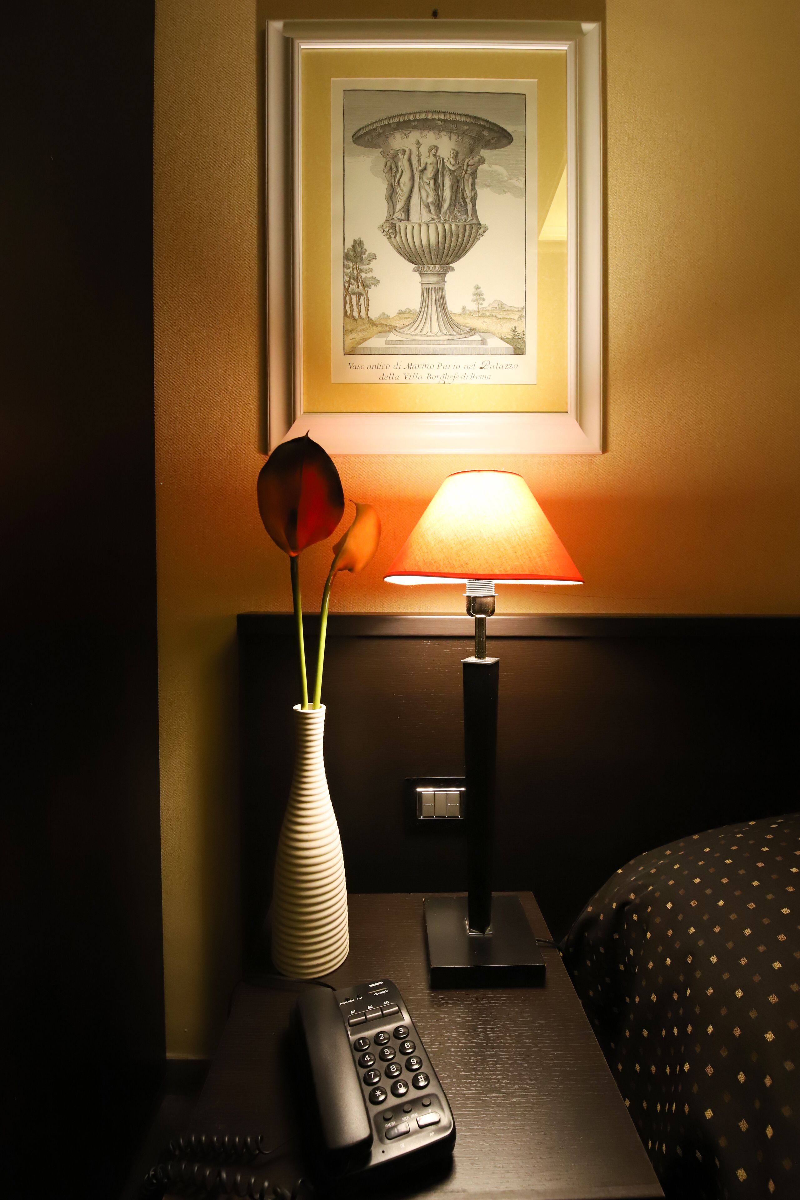 Ett hotellrum med ett inramat konstverk ovanför ett nattduksbord, en vas med en enda blomma, en lampa och en fjärrkontroll.
