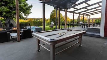 An outdoor pool table area with a covered seating area, surrounded by outdoor furniture and a view of a landscaped garden.